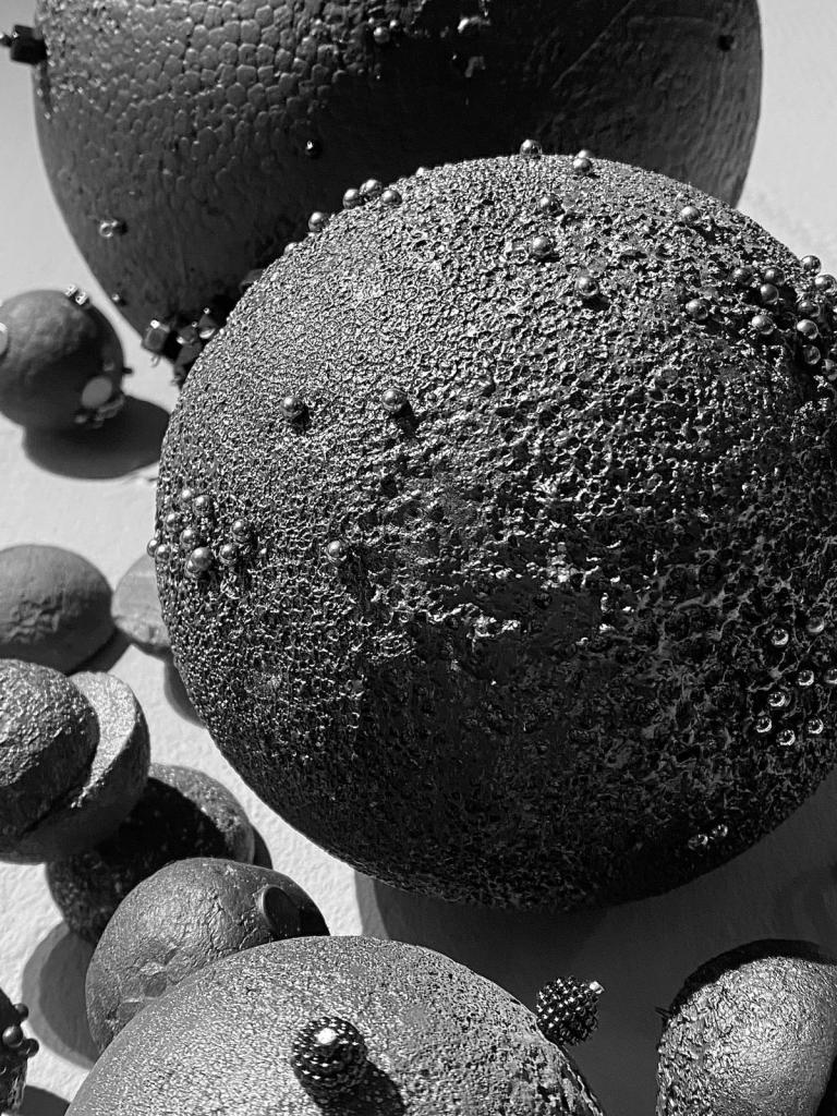 Roxana Donaldson, Saftey First art installation, Assemblage art black spheres, detail, 2025. Black porous sphere with pins, wires and nails on a white wall in official Safety First the Aftermath exhibtion, Bucharest, Galeria Calea Victoriei 91-93, 2025.